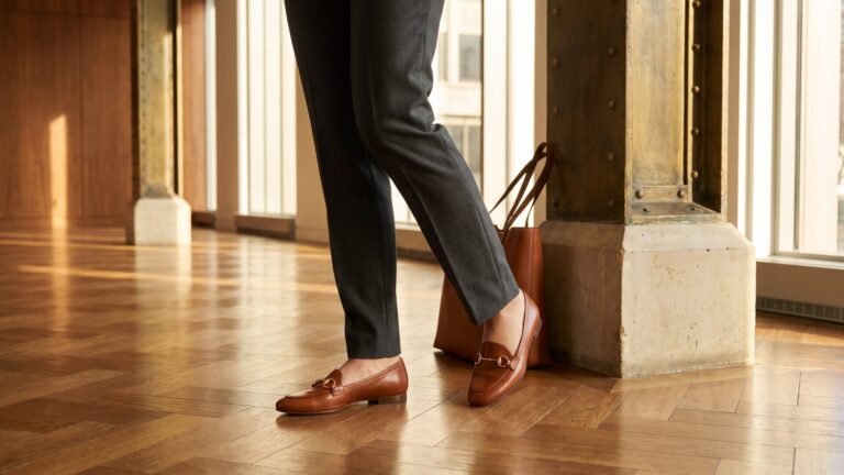 Person wearing brown leather horsebit flats and tailored trousers walking across a parquet wood office floor