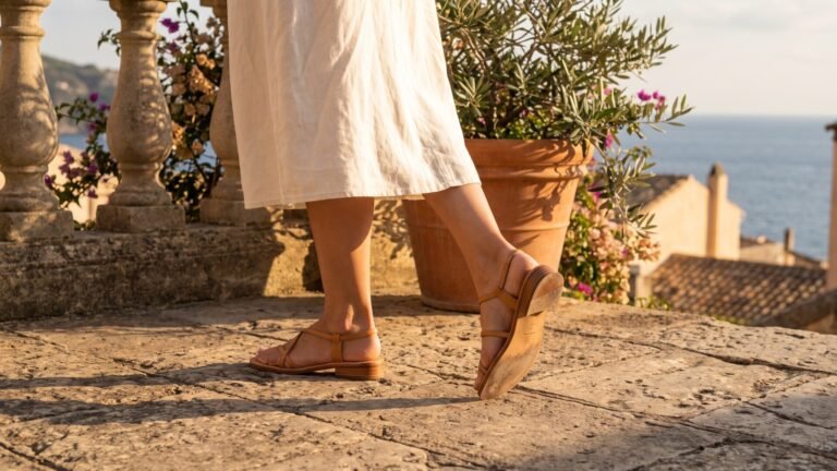 Woman walking across a stone terrace wearing tan leather strappy flat sandals and a white linen dress