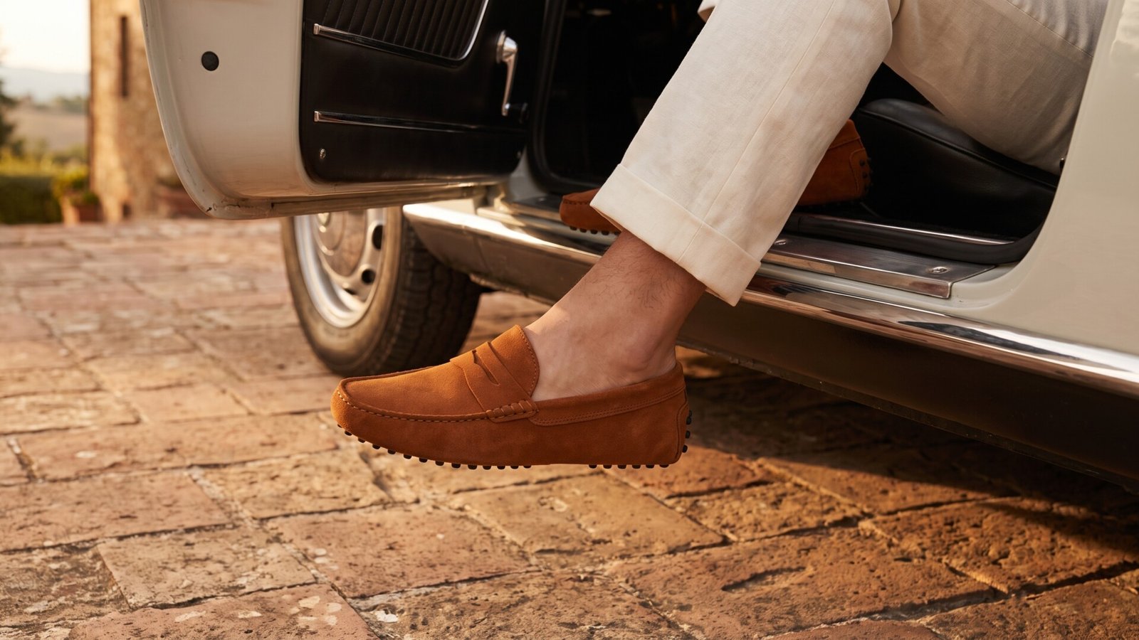 Brown suede Tod's Gommino driving loafers worn while stepping out of a vintage car onto cobblestone ground